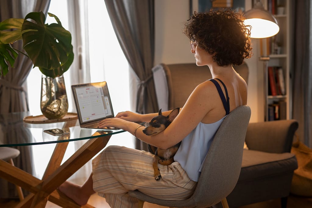 A person with curly hair sits at a glass table working on a laptop while holding a small dog in their lap.
