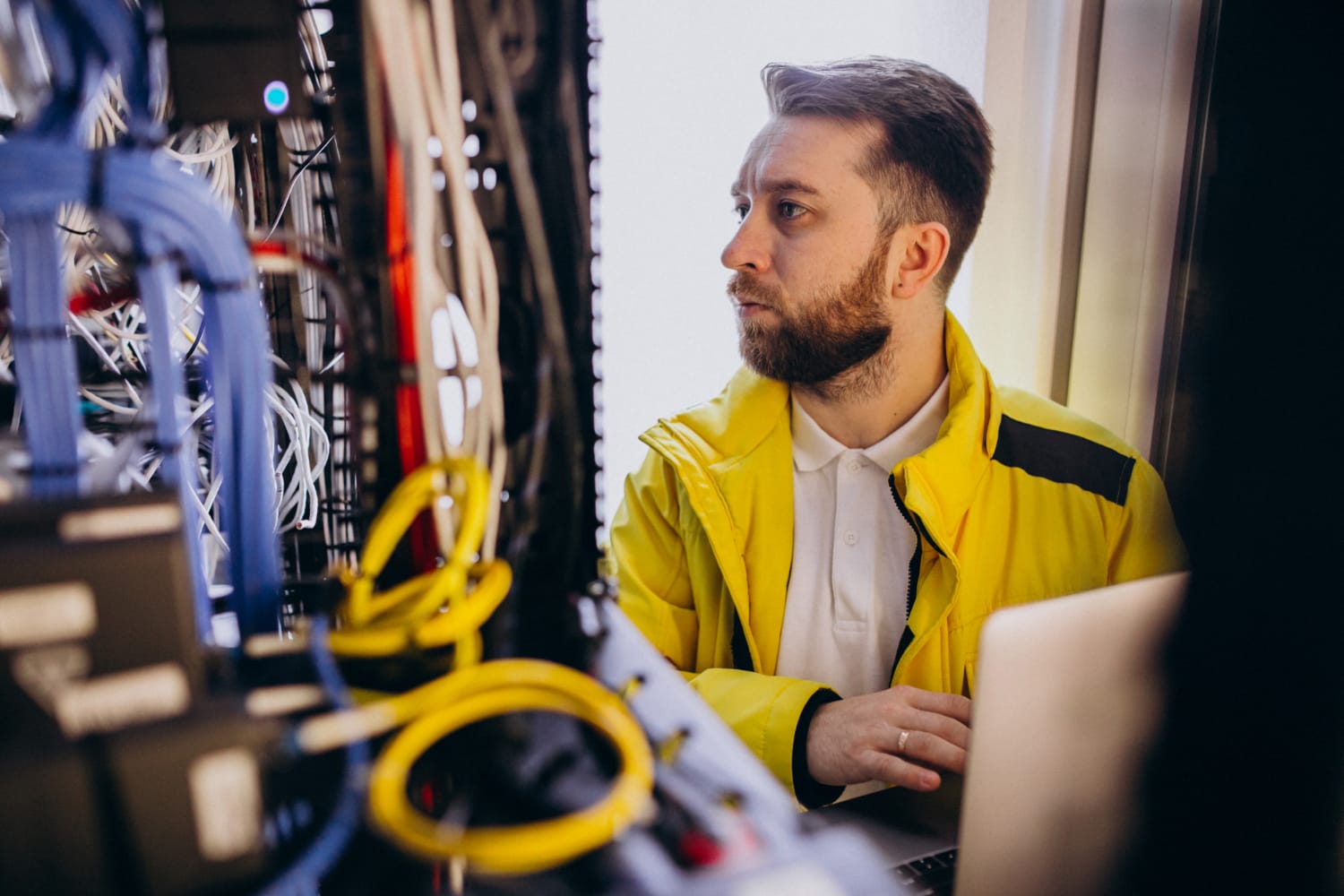 A technician in a yellow jacket works on a laptop in front of a server rack filled with cables.